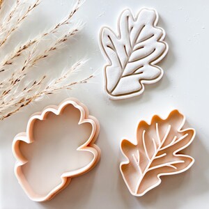 May include: A set of three light peach-colored plastic cookie cutters shaped like oak leaves. The set includes one solid outline cutter and two detailed stamp cutters that feature embossed vein patterns within the leaf shape for baking.