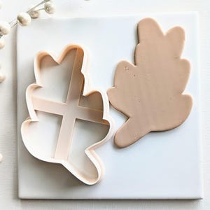 May include: A white plastic cookie cutter shaped like a leaf, next to a brown clay leaf.