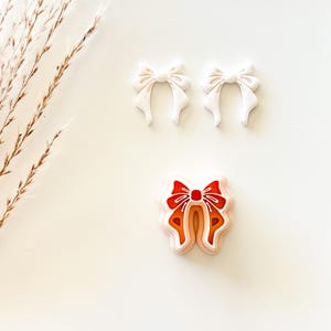 May include: Three bow-shaped cookie cutters. Two are solid white, and one is a layered design with red, orange, and peach tones. The cutters are arranged on a white surface, with a sprig of dried grass in the upper left corner.