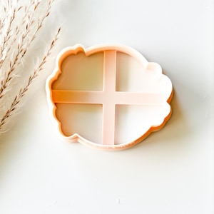 May include: A light peach-colored cookie cutter with a scalloped edge and a cross-shaped interior design. The cutter is made of plastic and is set against a white background. Dried ornamental grass is visible in the upper left corner.