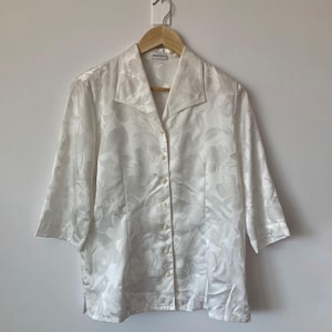 Könnte beinhalten: Ein weißes Hemd mit Knöpfen und einem Blumenmuster. Das Hemd hat einen Kragen und Dreiviertelärmel.