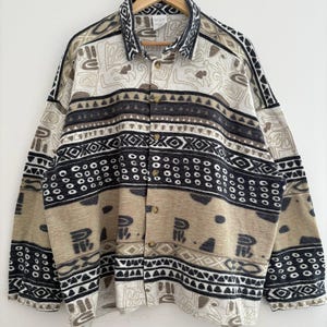 May include: A button-down shirt with a geometric pattern in beige, black, and white. The shirt is hanging on a wooden hanger against a white wall. The pattern includes various shapes and designs.