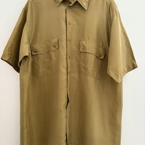 以下が含まれることがあります： 落ち着いたオリーブグリーンの半袖シャツ。ボタン留めの胸ポケットが2つと、クラシックな襟が付いています。木製のハンガーに掛けられており、背景は白です。