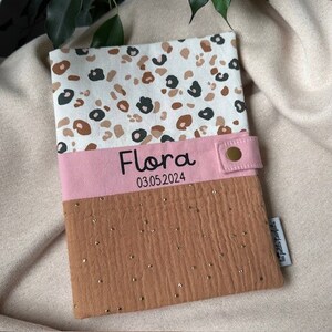 Peut inclure: Une couverture pour bébé rose et marron avec un motif léopard. La couverture a un ruban rose avec le nom "Flora" et la date "03.05.2024" imprimée dessus.