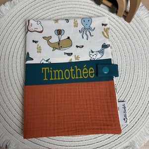 Peut inclure: Une bavette de bébé personnalisée avec une bordure vert sarcelle et un fond rouille. Le haut est en tissu blanc avec un imprimé d'animaux marins fantaisistes. Le nom "Timothée" est brodé en jaune sur la bordure vert sarcelle.