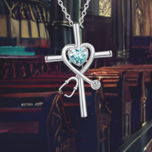 May include: Silver cross necklace featuring a blue heart and a stethoscope design. The heart has a clear blue gemstone at its centre.
