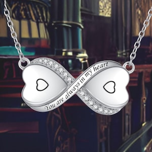 May include: Silver-toned necklace featuring an infinity symbol pendant. The pendant has two heart-shaped elements, each with a small heart outline. The infinity symbol is embellished with small, clear crystals and the inscription "You are always in my heart."