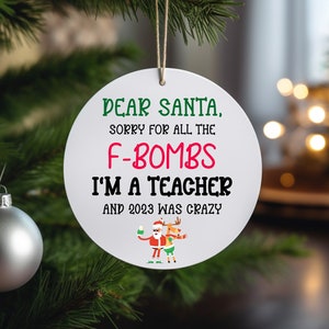 May include: White round Christmas ornament with a green, red, and white design. The ornament features the text "Dear Santa, Sorry for all the F-Bombs I'm a Teacher and 2023 was crazy". A cartoon image of Santa Claus and a reindeer are shown at the bottom of the ornament.