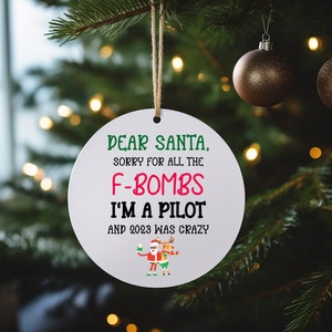 May include: White round Christmas ornament with the text "DEAR SANTA, SORRY FOR ALL THE F-BOMBS I'M A PILOT AND 2023 WAS CRAZY". A cartoon image of Santa Claus and a reindeer are at the bottom of the ornament.