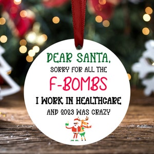 May include: White round Christmas ornament with the text "DEAR SANTA, SORRY FOR ALL THE F-BOMBS I WORK IN HEALTHCARE AND 2023 WAS CRAZY". The ornament features a cartoon image of Santa Claus and a reindeer.