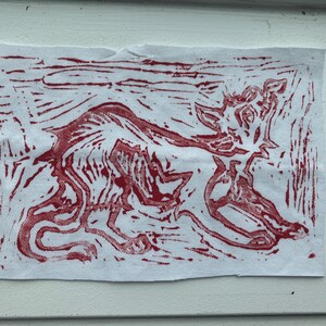 Puede incluir: Una pieza rectangular de tela blanca con una ilustración en impresión de bloque roja de un animal estilizado de cuatro patas en posición agachada. El diseño utiliza líneas audaces y texturizadas para representar la forma sobre el fondo blanco.