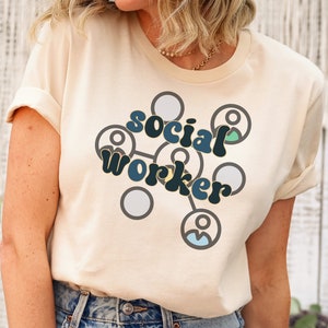May include: A woman wearing a light beige t-shirt with the text "social worker" in a retro font and a graphic of connected people. She is wearing blue jeans with distressed details.