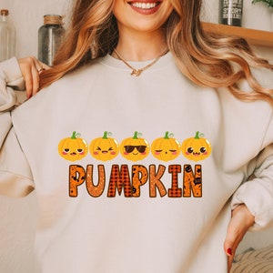 May include: A white sweatshirt with a graphic of five orange pumpkins with different facial expressions. The word "PUMPKIN" is written in orange letters with a black and orange plaid pattern.