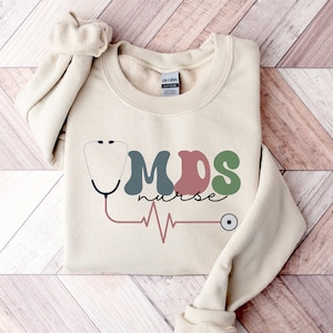 May include: A beige sweatshirt with the text "MDS nurse" in colorful letters, a stethoscope graphic, and a heartbeat line.