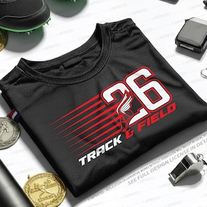 Op de afbeelding: Zwart atletisch t-shirt met het nummer 26 en de woorden "Track & Field" in wit en rood. Het shirt heeft een afbeelding van een gevleugelde voet. Medailles, een fluitje en een hardloopschoen zijn ook zichtbaar.