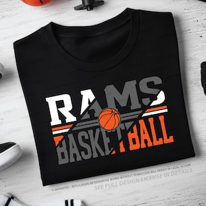 May include: Black t-shirt with the words "RAMS BASKETBALL" in white, gray, and orange. An orange basketball graphic is centered in the design. The shirt is displayed on a light-colored surface with athletic accessories.