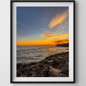 May include: A framed photograph of a rocky coastline with a sunset over the ocean. The sky is a mix of blue, orange, and pink, with clouds reflecting the colors of the sunset. The water is calm and reflects the sky.