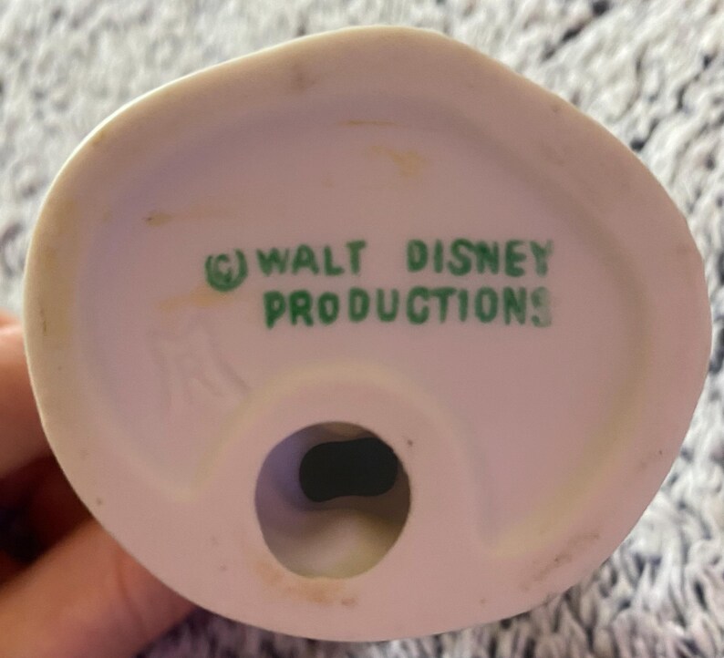 May include: A close-up of the bottom of a white ceramic figurine. The text "(C) WALT DISNEY PRODUCTIONS" is printed in green. The figurine has a circular hole at the base.