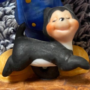 May include: A ceramic figurine of a black and white cat with a smiling face and closed eyes. The cat is posed on a yellow and brown base, with a blue and yellow figure in the background.