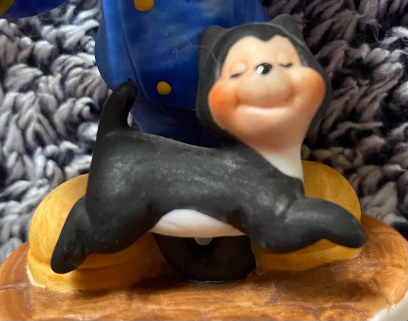 May include: A ceramic figurine of a black and white cat with a smiling face and closed eyes. The cat is posed on a yellow and brown base, with a blue and yellow figure in the background.