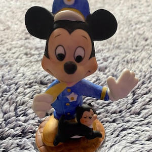May include: A ceramic figurine of Mickey Mouse in a police uniform, with a small black cat at his feet. Mickey is wearing a blue uniform with a yellow stripe and a police hat. The cat is black with a smiling face. The figurine is on a brown base.