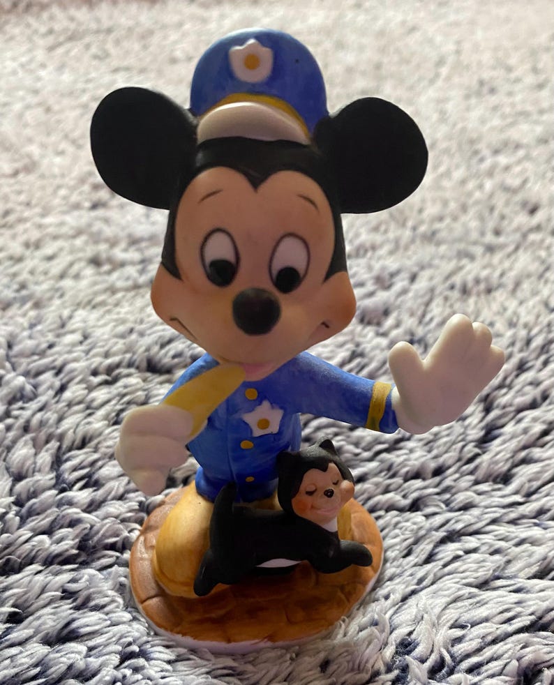 May include: A ceramic figurine of Mickey Mouse in a police uniform, with a small black cat at his feet. Mickey is wearing a blue uniform with a yellow stripe and a police hat. The cat is black with a smiling face. The figurine is on a brown base.