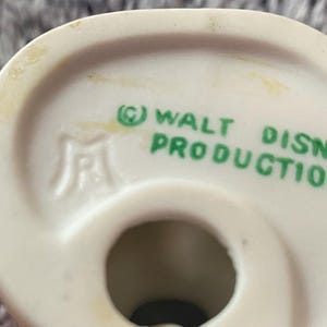 May include: Close-up of a white ceramic figurine base with the text "&copy; WALT DISNEY PRODUCTIONS" in green. The figurine has a hole in the center and a logo on the side. The background is a blurred, light gray surface.