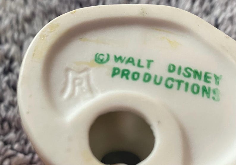 May include: Close-up of a white ceramic figurine base with the text "&copy; WALT DISNEY PRODUCTIONS" in green. The figurine has a hole in the center and a logo on the side. The background is a blurred, light gray surface.