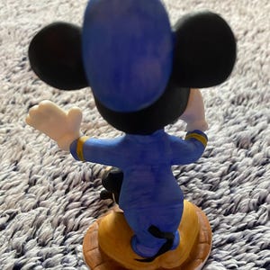 May include: A ceramic figurine of Mickey Mouse from the back, wearing a blue uniform with gold accents. The figure has black ears and a yellow base with a brown border. Mickey's white gloved hand is raised.