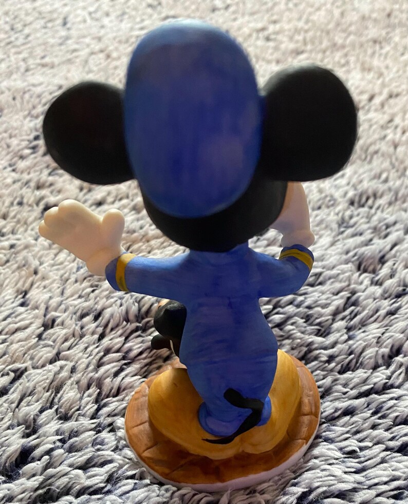 May include: A ceramic figurine of Mickey Mouse from the back, wearing a blue uniform with gold accents. The figure has black ears and a yellow base with a brown border. Mickey's white gloved hand is raised.