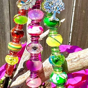May include: A colorful beaded suncatcher with green, pink, and red beads, and silver charms of a sunflower, a leaf, and a tree. The suncatcher is hanging from a black metal hook.