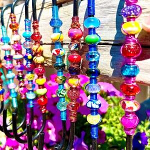 May include: A set of colorful beaded hooks made with wire and glass beads. The hooks are black metal and are hanging from a wooden surface. The beads are in various colors, including red, blue, green, yellow, and purple.