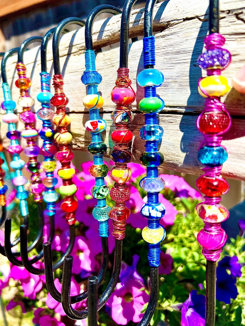 May include: A set of colorful beaded hooks made with wire and glass beads. The hooks are black metal and are hanging from a wooden surface. The beads are in various colors, including red, blue, green, yellow, and purple.