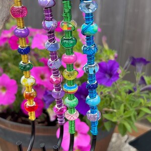 May include: Five colorful beaded hooks made of wire wrapped around metal hooks. The beads are various colors, including purple, green, blue, and yellow. The hooks are hanging from a branch.