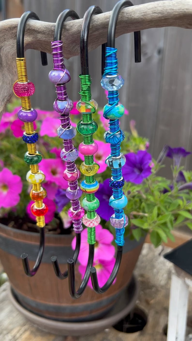 May include: Five colorful beaded hooks made of wire wrapped around metal hooks. The beads are various colors, including purple, green, blue, and yellow. The hooks are hanging from a branch.