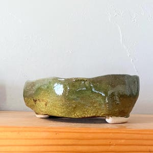 May include: A handmade, low-profile ceramic bowl with a textured, glazed surface. The bowl is primarily olive green with hints of gold and sits on three small, white feet. The rim has an uneven, organic shape, suggesting a handcrafted origin.