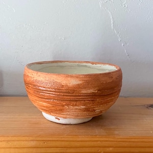 May include: A handmade ceramic bowl with a textured, reddish-brown exterior and a white base. The bowl has a wide opening and a smooth, off-white interior. The bowl is sitting on a wooden surface.