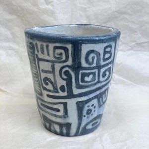 May include: A white ceramic cup with a blue geometric pattern. The pattern is made up of squares, circles, and lines.