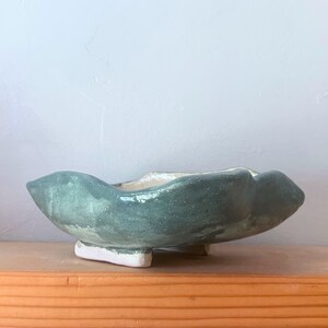 May include: A shallow, hand-formed ceramic bowl with an irregular, wavy rim. The exterior features a muted, dusty blue-green glaze with visible texture, while the interior and the small, blocky feet at the base are finished in a contrasting white.
