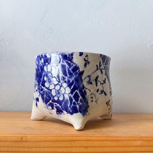 May include: A handcrafted, ceramic planter with a white base and blue floral patterns. The planter has a square shape with three small feet. The blue design features a combination of floral and geometric elements, creating a unique aesthetic.