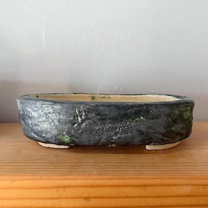 May include: A handmade, oval-shaped bonsai pot. The exterior is a textured, dark charcoal color with hints of green. The interior is a light beige. The pot is elevated on small, rectangular feet, and is suitable for small plants.