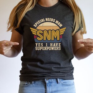 May include: Dark gray t-shirt with a retro design. The graphic reads "SPECIAL NEEDS MOM SNM YES I HAVE SUPERPOWERS" in a vintage style with wings and a sunset background. The shirt is made of a soft material.
