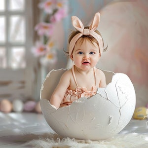 Peut inclure: Un bébé portant un bandeau de lapin rose est assis dans un œuf blanc fissuré en guise d'accessoire. Le bébé porte une robe rose et a les yeux bleus. L'arrière-plan est rose clair et blanc.