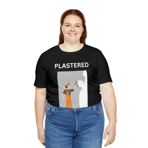 Puede incluir: Una camiseta negra con un gráfico blanco de una persona enyesando una pared. El texto "PLASTERED" está por encima del gráfico.
