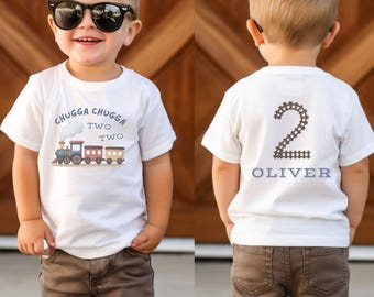 May include: A white short-sleeved t-shirt featuring a graphic of a train with the text Chugga Chugga Two Two on the front. The back of the shirt displays the number 2 formed by train tracks above the name Oliver in blue block lettering.