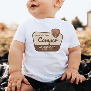 May include: White baby onesie with a brown patch that says "One Happy Camper Adventuring Since 2024" with a small camping scene inside a brown circle.