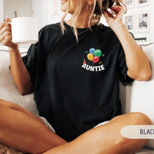 K&ouml;nnte beinhalten: Schwarzes T-Shirt mit einem bunten Ballon-Motiv und dem Wort "AUNTIE" in Wei&szlig;. Die Ballons sind blau, gr&uuml;n, gelb und rot. Das Shirt hat einen l&auml;ssigen Stil.