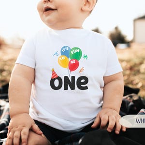 K&ouml;nnte beinhalten: Ein wei&szlig;es T-Shirt mit einer bunten Grafik zum ersten Geburtstag. Das Design umfasst Luftballons mit den Worten "It's Fun To Be One" und eine Partyh&uuml;tchen. Das Wort "ONE" steht in gro&szlig;en, fetten, schwarzen Buchstaben.
