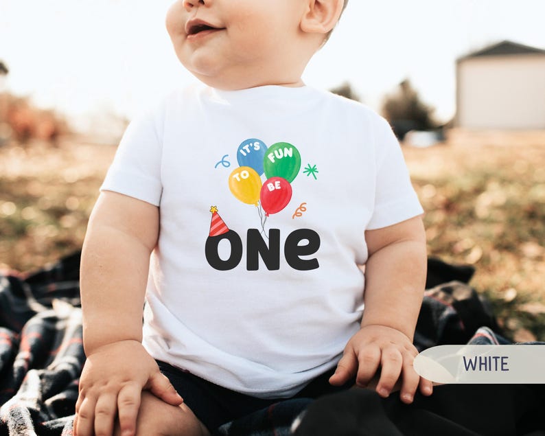 K&ouml;nnte beinhalten: Ein wei&szlig;es T-Shirt mit einer bunten Grafik zum ersten Geburtstag. Das Design umfasst Luftballons mit den Worten "It's Fun To Be One" und eine Partyh&uuml;tchen. Das Wort "ONE" steht in gro&szlig;en, fetten, schwarzen Buchstaben.