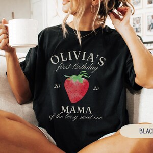May include: Black t-shirt with the text "OLIVIA'S first birthday" above a red strawberry graphic. Below the strawberry is the text "MAMA of the berry sweet one". The numbers "20" and "25" are on either side of the strawberry.